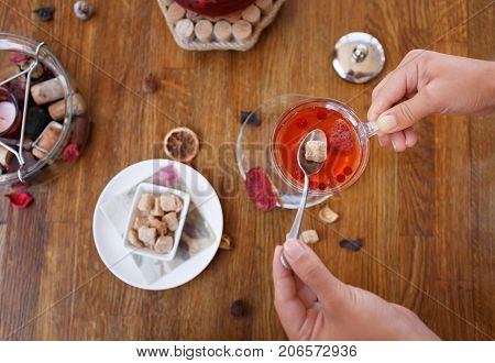 Top view of a bowl of crunchy biscuits with nuts and spices on a wooden background. Woman's hands holding a spoon with brown sugar and a cup of red, hot strawberry tea. A big and full tea kettle.