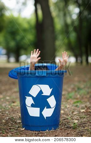 Baby's hands, sticking out from the blue bin. Playing with the trash bin, in the street, summer outdoor, save the planet, save the Earth concept. Day in park. Funny babies.