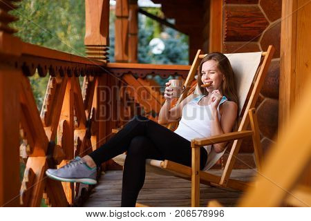Young attractive woman sitting outdoors on deck chair with coffee cup and oat cookie at mountain resort. Female on chaise-longue with tea mug and snack. Diet concept. Green forest on background.