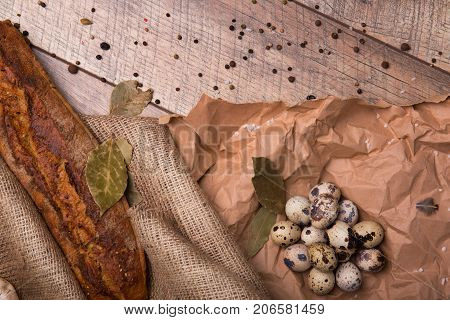 Top view of brown rye bread, speckled fresh quail eggs, dried bay leaves, grocery paper and different seasonings on a wooden background.
