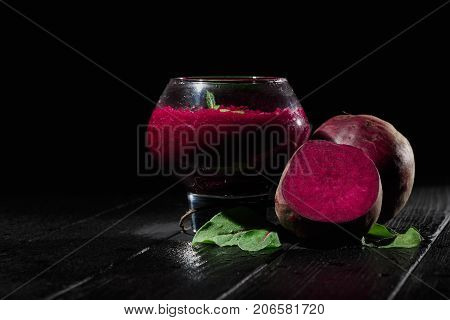 A big, fancy glass filled with saturated red beetroot juice on a black wooden background. Ripe and peeled beets for a healthful summer salad. Raw vegetable cocktails. Copy space.