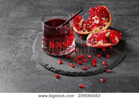 A transparent glass full of sweet pomegranate juice and a cut garnet on a round plate on a dark gray background. Summer, healthful and fruity red cocktail with black straw and fresh garnet.