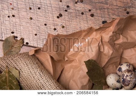 Closeup of a sheet of grocery paper, speckled quail eggs, dried up aromatic bay leaves and different seasonings, wooden table on a light wooden background.