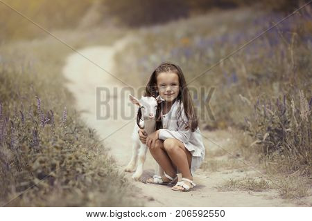 Little girl plays and huhs goatling in country, spring or summer nature outdoor. Cute kid with baby animal, forest, trod, glade background. Friendship of child and yeanling, image toned.