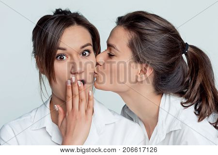 Studio portrait of young female twins sisters on gray. The one girl kissing another on the cheek