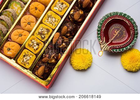 stock photo of Indian sweet or mithai and oil lamp or diya with gift box and flowers on decorative or colourful background, selective focus