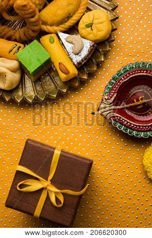 stock photo of Indian sweet or mithai and oil lamp or diya with gift box and flowers on decorative or colourful background, selective focus