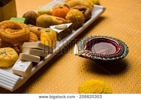 stock photo of Indian sweet or mithai and oil lamp or diya with gift box and flowers on decorative or colourful background, selective focus
