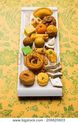 stock photo of Indian sweet or mithai and oil lamp or diya with gift box and flowers on decorative or colourful background, selective focus