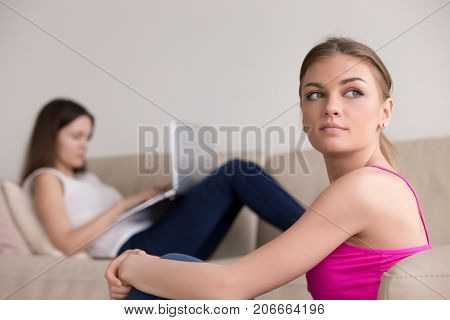 Candid portrait of attractive thoughtful girl and girlfriend typing on laptop on background. Dreamy woman thinking about life while her friend busy on computer. Bored young pretty lady daydreaming.