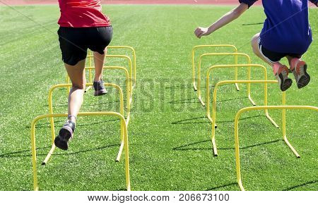 Two athletes perform coordination and strength drills by jumping over mini hurdles on a green turf field at practice.