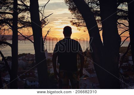 Sunset view of male silhouette admiring popular touristic destination from the top of hill. Panoramic view of beautiful Croatian landscape with rolling hills gardens and houses of the ancient town.