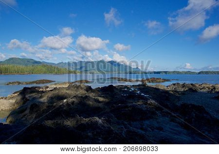 Shadows partially obscure the rocks on the shore of Battle Bay on a beautiful afternoon west caost of Vancouver Island. The sky is blue and fluffy clouds float above the distant mountains.