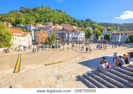 Sintra, Portugal - August 8, 2017: Tourists sitting on steps of National Palace of Sintra in the popular main Amelia Square with Castle of the Moors on background. Sintra urban center in a sunny day.