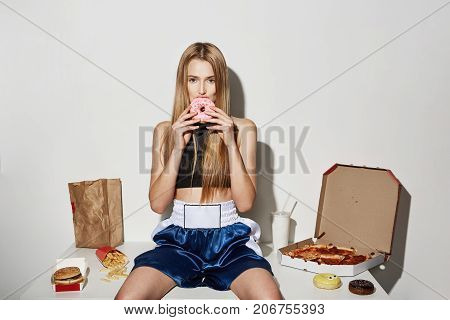 Joyful beautiful female model with blonde hair in sport wear sitting on table, eating junk food, looking in camera with cute donut in mouth.