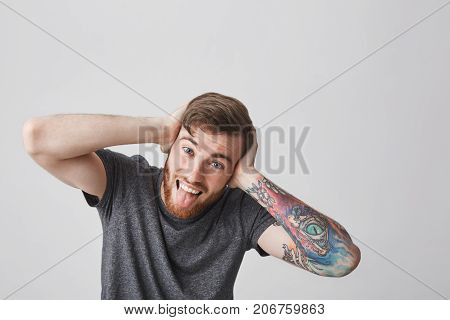 We're all mad here. Portrait of good-looking funny european man with beard and tattooed arm looking in camera with crazy expression, clothing ears with hands, showing tongue and having fun