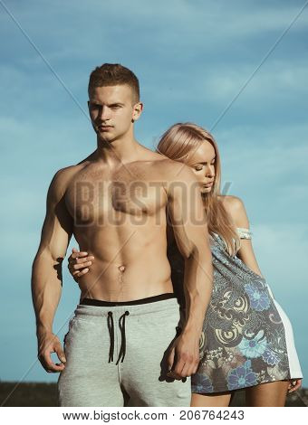 Girl with blond hair hugging bodybuilder with muscular torso. Couple in love on sunny summer day. Bodybuilding and fitness. Man and woman posing on blue sky. Vacation and lifestyle concept