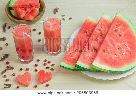Two hearts, watermelon juice into two glass cups with a straw on a light wooden background, a delicious cocktail, a plate with slices of ripe watermelon on the table. Red berries, cranberries.