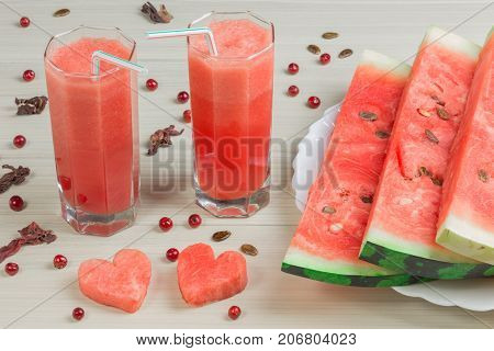 Two hearts, watermelon juice into two glass cups with a straw on a light wooden background, a delicious cocktail, a plate with slices of ripe watermelon on the table. Red berries, cranberries.