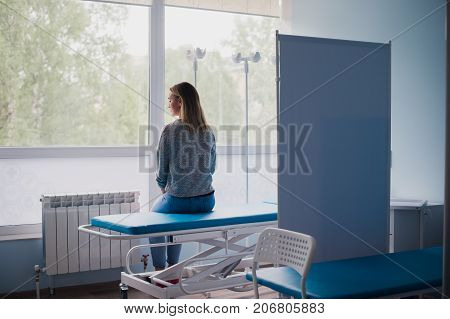 Young pregnant woman sitting on bed in comfortable ward, waiting for doctor thoughtfully.