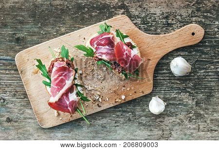 Brushetta set with smoked meat, arugula, garlic and dried tomatoes on a rustic wooden board over a rough wood background. Top view