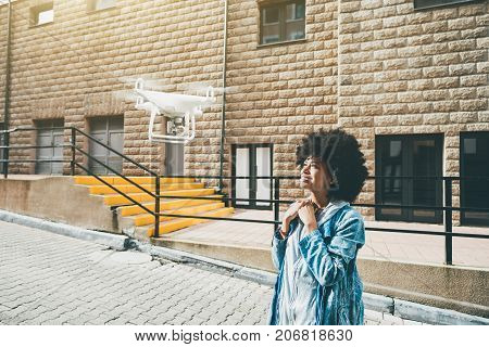 Smiling cute black girl is recording her video blog using modern flying drone: she is standing in the center of urban street and looking up into camera of quadrupter with facade of building behind