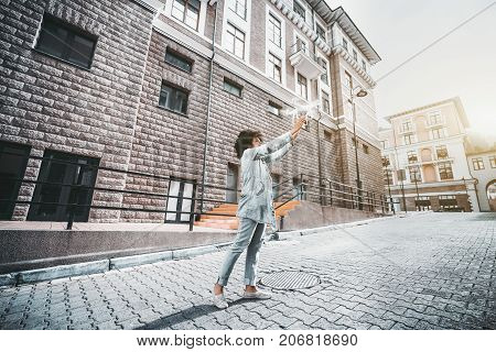 Curly young african-american woman in jean jacket is standing on pavement in the middle of urban street and launching white working quadrupter; sunny autumn day wide-angle shooting