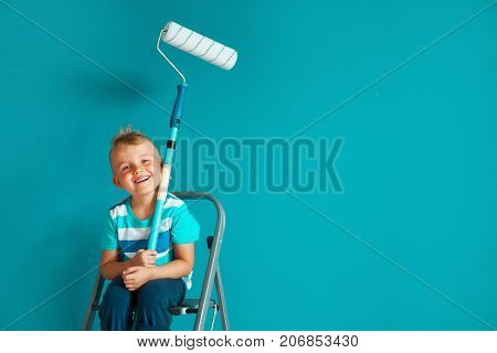 The Child Paints The Blue Wall With A Roller. The Boy Holds A Large Brush For Painting. Home Repairs