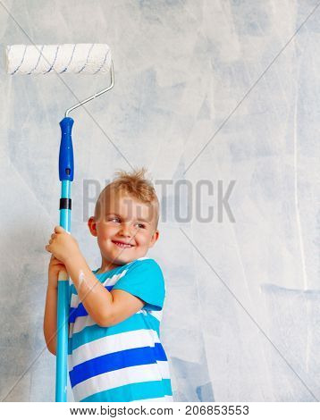 The Child Paints The Blue Wall With A Roller. The Boy Holds A Large Brush For Painting. Home Repairs