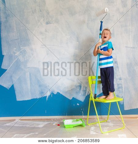 The Child Paints The Blue Wall With A Roller. The Boy Holds A Large Brush For Painting. Home Repairs