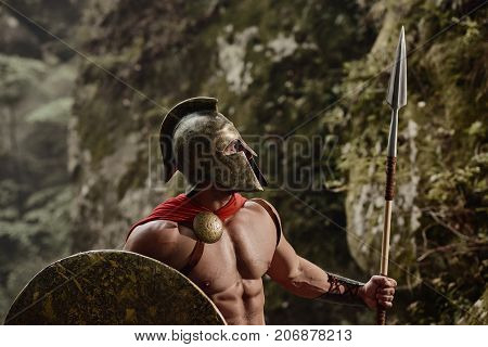 Strong muscular Roman gladiator in helmet and red cloak posing shirtless with shield and spear looking confidently away. Spartan.