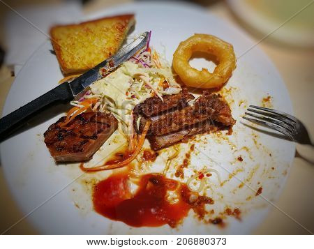 Leftover of pepper pork steak on white plate, including onion ring, bread, and side salad (slided orange carrot, green and purple cabbages); metal knife and fork on dish; selective focus with vignette; concept of organic waste
