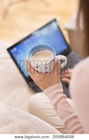 Detail of a young woman working on her laptop and drinking coffee. Selective focus on the coffee cup