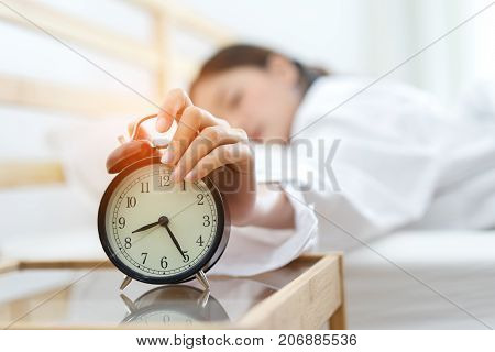 Young asian woman press the button snooze wake up alarm clock alert in the morning and sleeping lazy next on bedroom at home