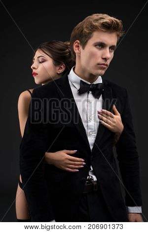 Sexy gentleman in a siut and bow-tie with his girl-friend embracing him cropped isolated shot on black background