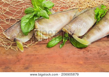 Several uncooked carcasses of the notothenia fish without of a heads and tails and twigs of basil and parsley closeup on the fishing net on a dark wooden surface