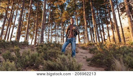 Portrait of a smiling young African man wearing a backpack standing alone on a trail while hiking in the forest on a sunny day