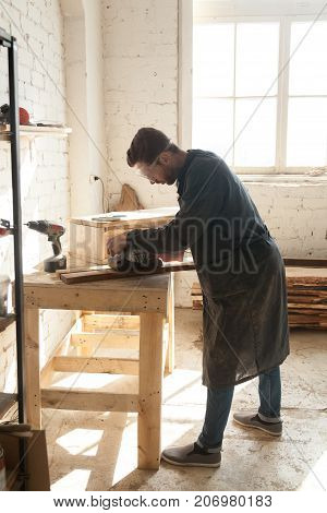 Carpenter polishes wooden boards with belt sanding machine while working in workshop. Young man choosing working profession at time of shortage skilled labor trades. Male millennial first job concept