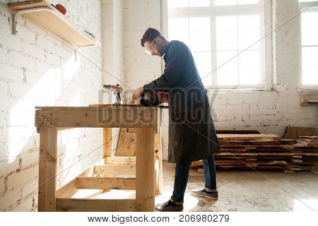 Joiner using electric tools while working in carpentry. Millennial worker getting on-the-job trailing in woodworking workshop, improve his skills in profession. Short-term job opportunities concept