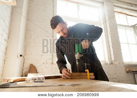 Skilled professional carpenter in workwear using electric drill machine, custom woodworking, making woodware, young experienced craftsman drilling wooden planks on table at workshop. Small business
