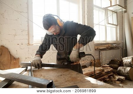 Young workman in protective equipment using angle grinder to smooth metal profile pipe in workshop interior with wooden planks and lumber. Focused on work skilled worker grinds steel parts with sparks