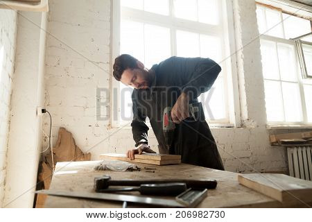 Joiner drilling wooden boards on workbench with electric drill. Young carpenter makes custom cabinet, repairs antiquarian furniture, working in workshop. Part time small business opportunity concept