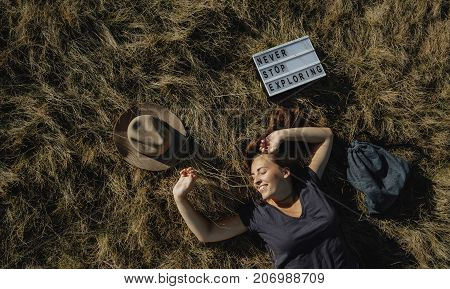 Top View Young Woman Traveler Lies On Dry Grass, Hat, Backpack, Whiteboard With Text Never Stop Expl