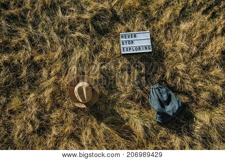 Flat lay travel composition with hat backpack board with text Never stop exploring on dry grass Top view