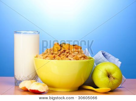 leckere Cornflakes in grüner Schale, Äpfel und Glas Milch auf Holztisch auf blauem Hintergrund