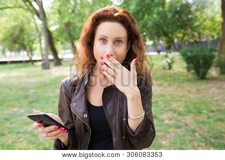 Embarrassed Lady Holding Smartphone And Covering Mouse In Park. Pretty Young Woman Looking At Camera