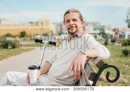 An Attractive Elderly 50-year-old Man With Long Hair In The Tail Sits In A Public Garden On A Bench.