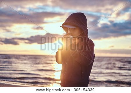 Cute Little Kid Is Enjoying Hot Beverege In Cold Evening At Seaside. There Are Beautiful Sunset Behi