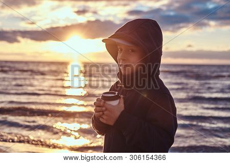 Cute Little Kid Is Enjoying Hot Beverege In Cold Evening At Seaside. There Are Beautiful Sunset Behi