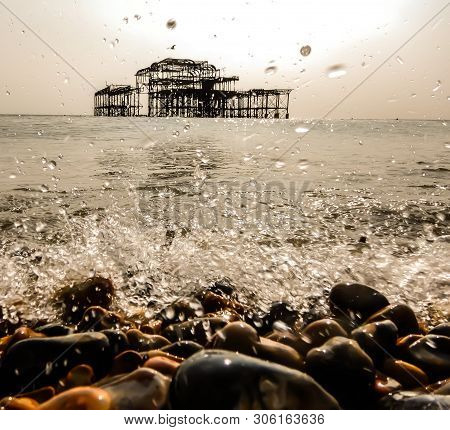 Splashing Water On Rocky Beach In Foreground With An Abandoned Old Pier On The Horizon In The Backgr